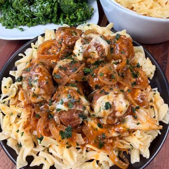 Pasta üzerinde et köfteleri, soğan ve sos ile servis edilen bir yemek, yan tarafta brokoli ve bir kase makarna yer alıyor.